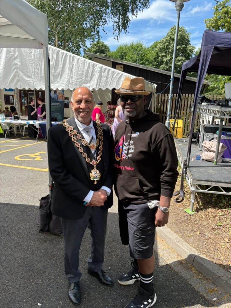 Two men smiling at community event, one in formal suit and chain, the other in casual hoodie and hat.