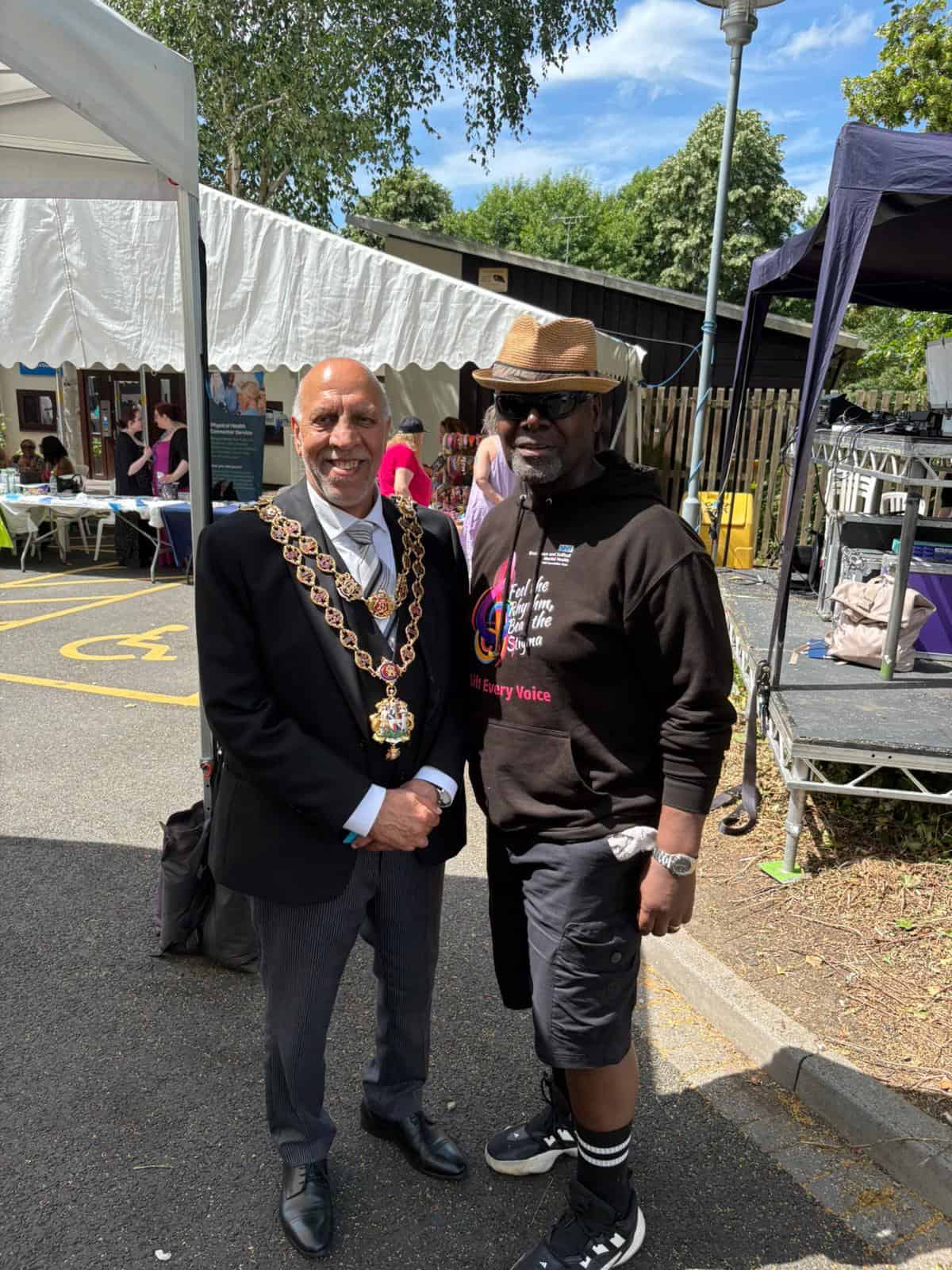 Two men smiling at community event, one in formal suit and chain, the other in casual hoodie and hat.